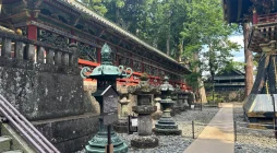 Camino de piedra en Nikkō con linternas de bronce, rodeado de templos tradicionales y naturaleza.